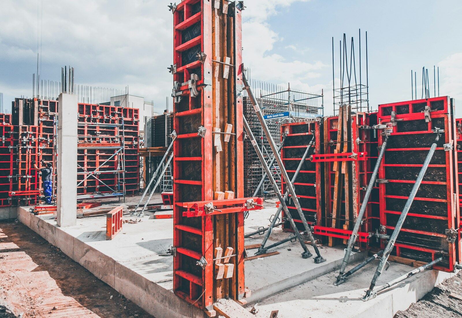 Czerwone stalowe elementy szalunku ściennego ze stemplami rektyfikacyjnymi stoją na świeżo zabetonowanej płycie fundamentowej.
